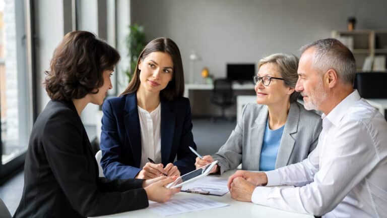 Four professionals, three women and one man, sit at a table in an office discussing documents. One woman holds a tablet as they engage in a formal business meeting focused on financial education and retirement income strategies.