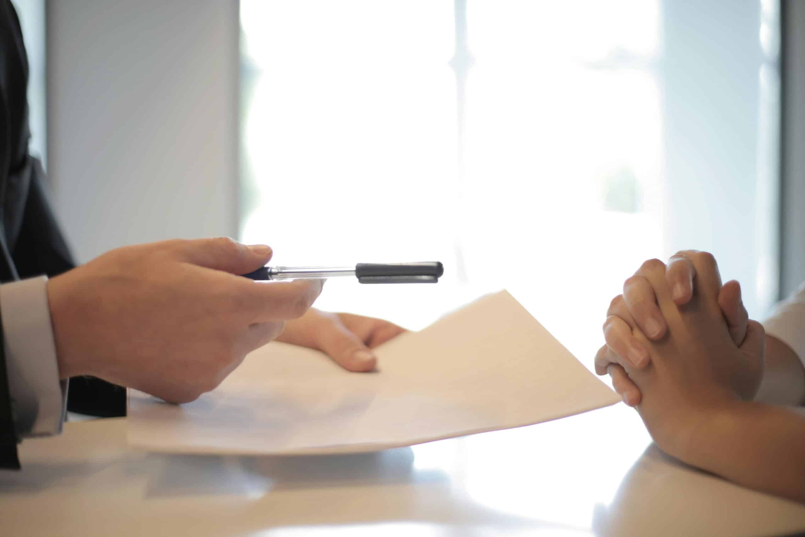 A person in a suit hands a pen and document to another person sitting across a table, suggesting a signing or agreement process related to mortgage services in a bright, professional setting.
