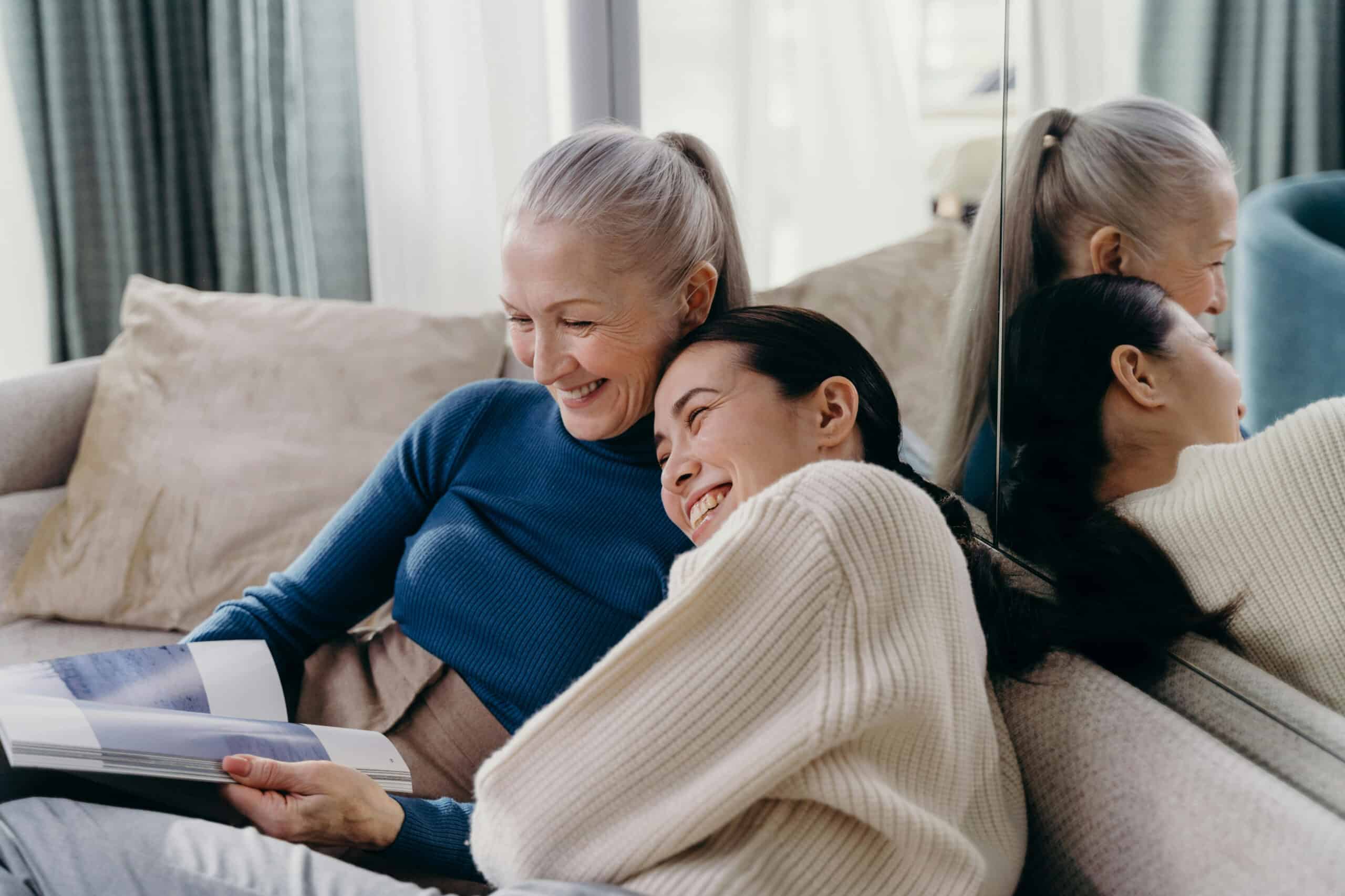 Two women sit closely on a couch, smiling and reading a book on financial education together. One has gray hair and wears a blue top; the other has dark hair and wears a cream sweater. They appear happy and relaxed, sharing a warm moment.