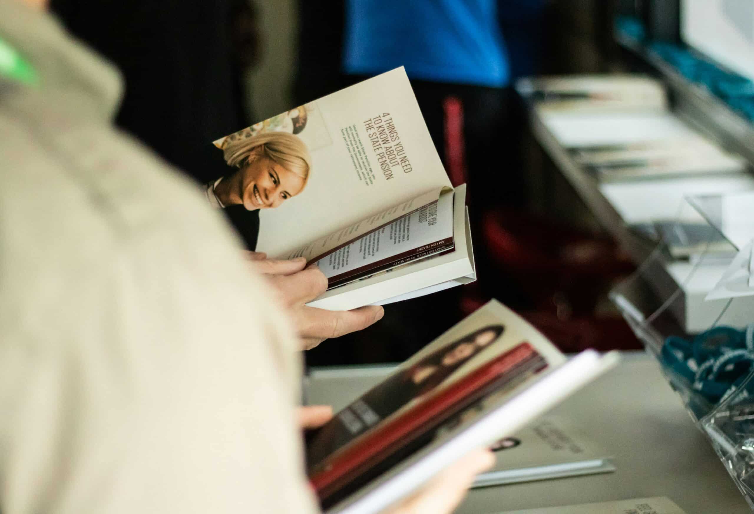 A person holds open a book displaying a smiling woman’s photo and text on financial education, while another browses a different book nearby on a table with more books and items.