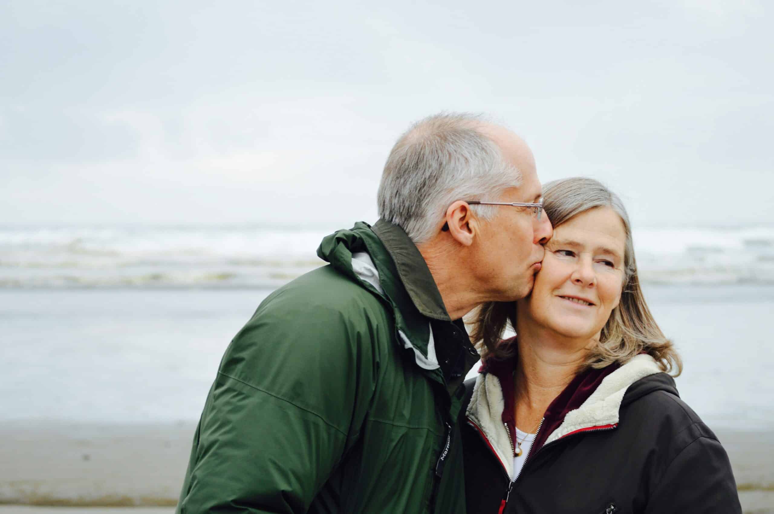 An older man kisses a smiling woman on the cheek while standing together on a beach, both wearing jackets. The ocean and cloudy sky are visible in the background, reflecting their peace of mind from smart retirement income strategies.