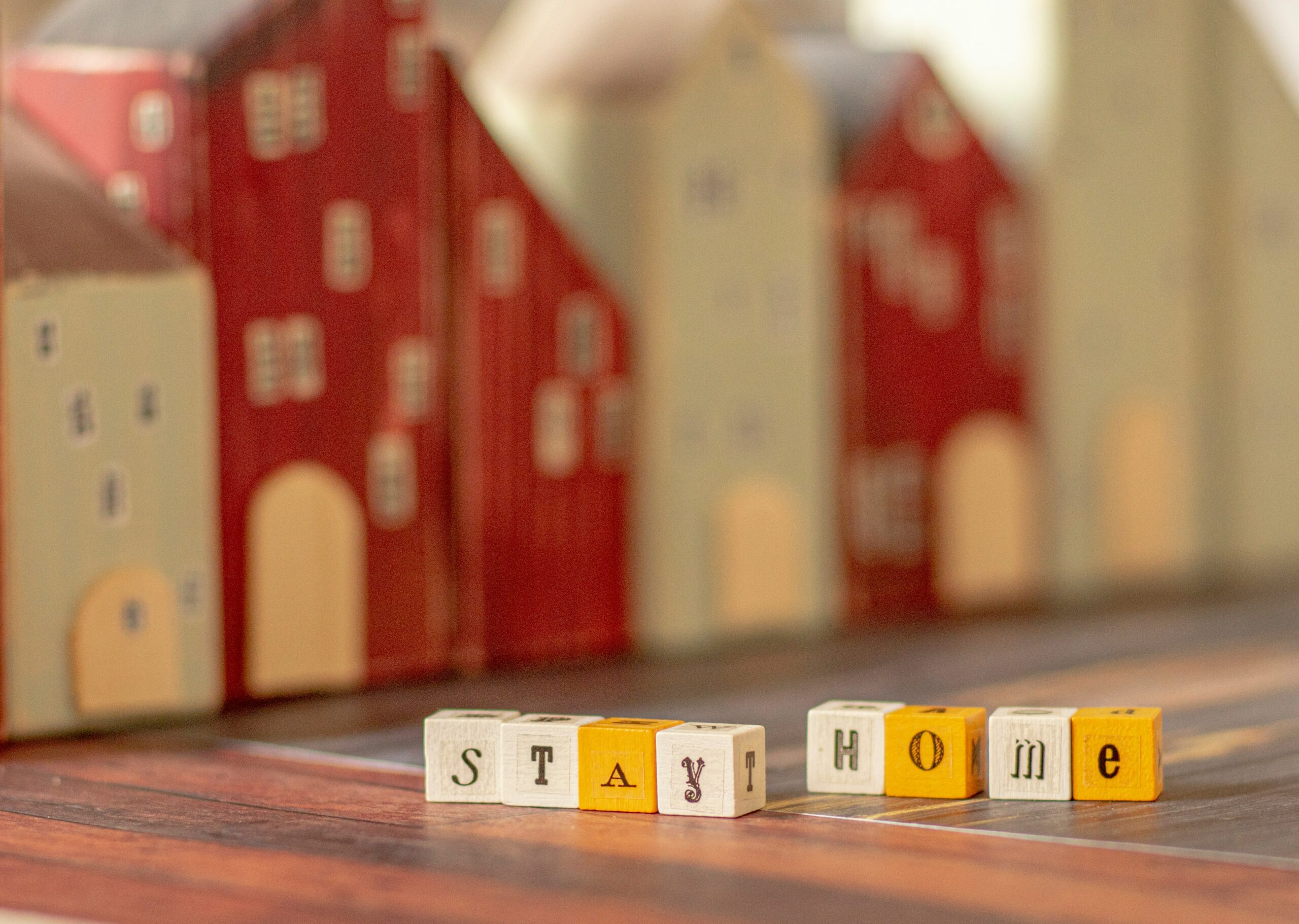 Wooden blocks spelling STAY HOME are arranged on a table, with blurred miniature wooden houses in the background. The scene conveys a cozy atmosphere, perfect for learning about mortgage services or exploring retirement income strategies.