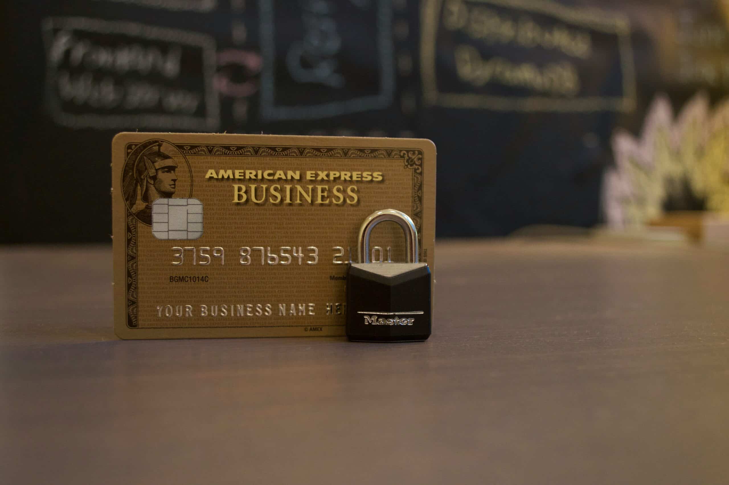 A gold American Express business credit card stands upright on a table, with a small black padlock in front; a blurred chalkboard in the background hints at financial education and smart retirement income strategies.
