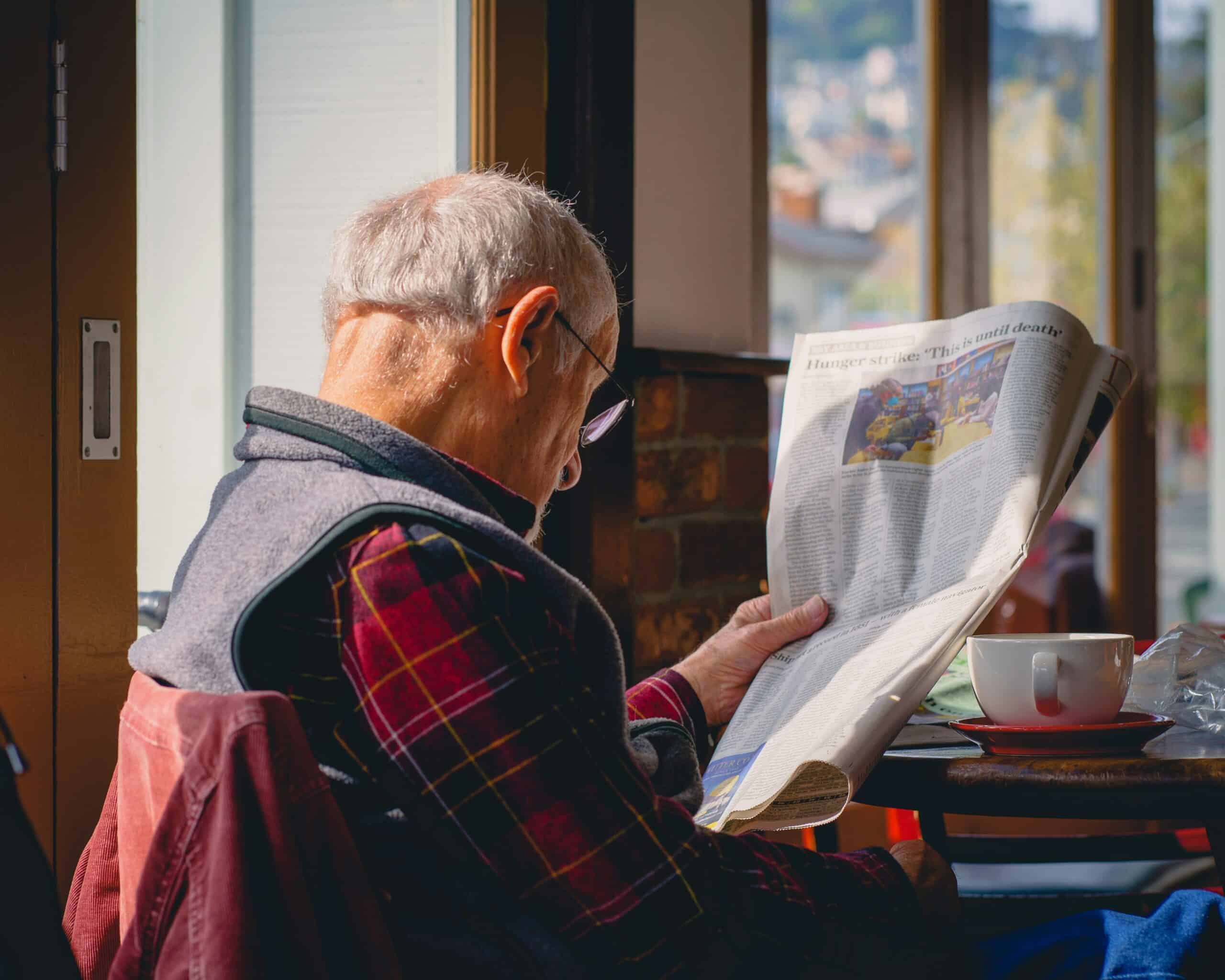 An elderly man with glasses sits by a window in a cozy cafe, reading about retirement income strategies. Sunlight streams in, illuminating his face and a white coffee cup on the table beside him.