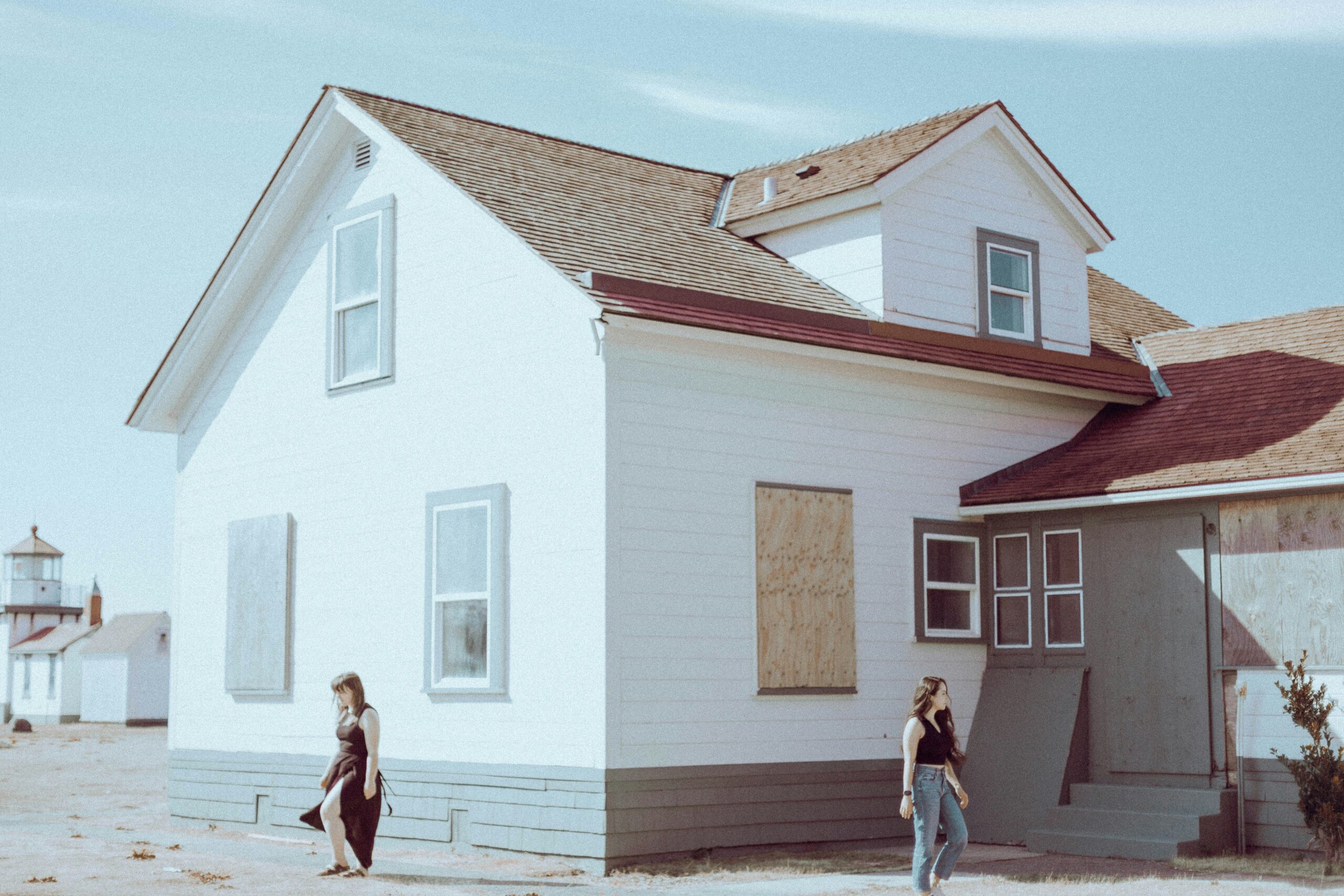 Two people walk past a white, boarded-up house under a blue sky. The calm scene hints at stories of mortgage services and financial education, while another building stands quietly in the background.