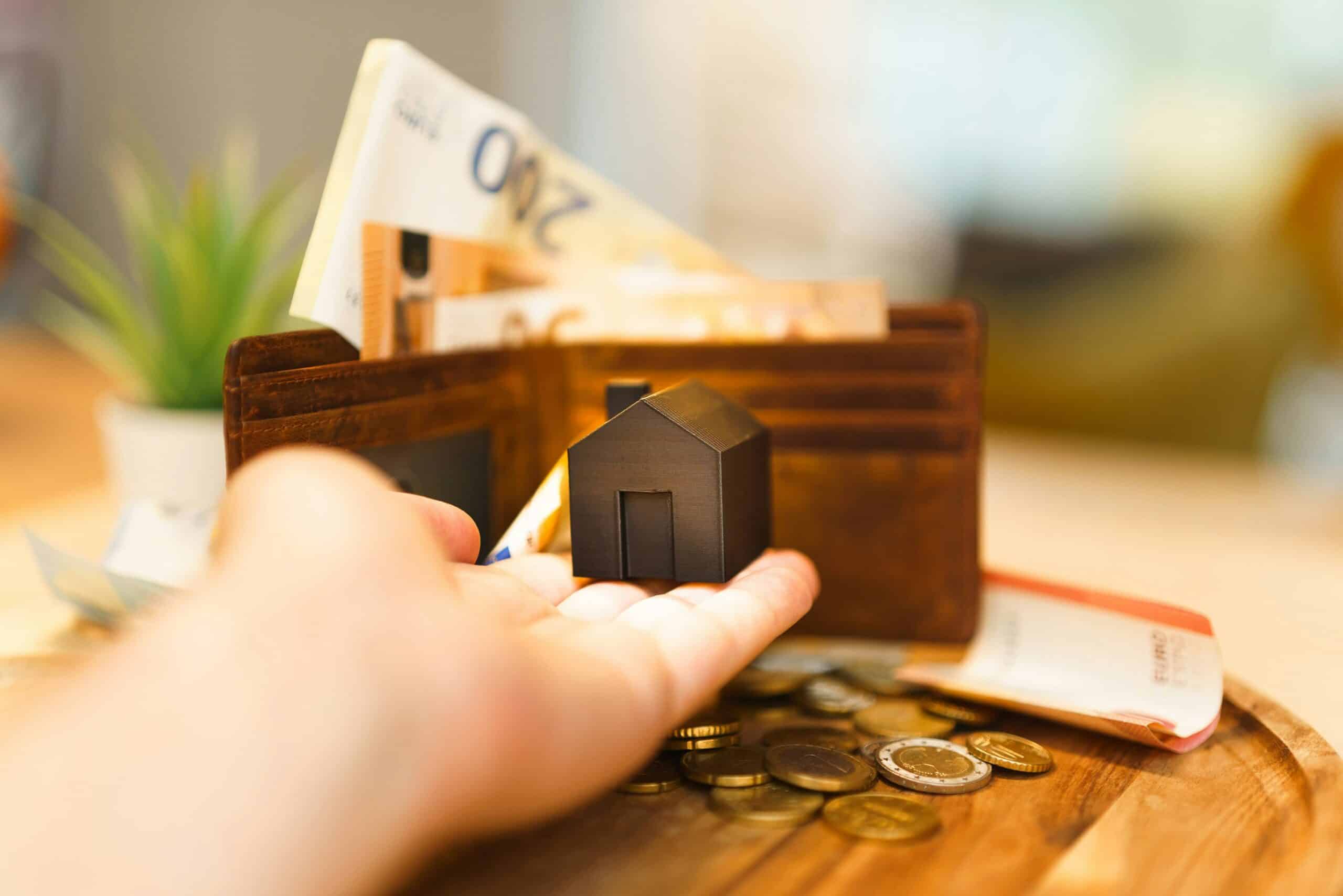 A hand holds a small house model in front of an open wallet with euro banknotes and coins scattered on a table, illustrating themes of home finance, mortgage services, or real estate investment.