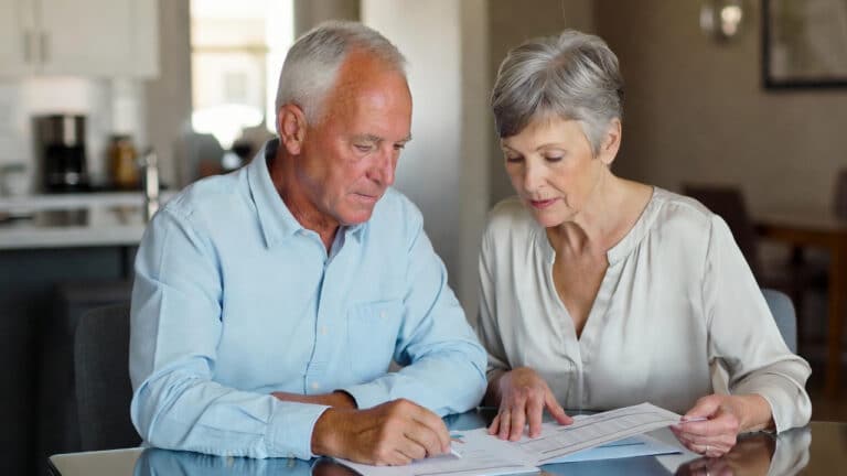 An older man and woman sit at a table reviewing documents together in a bright kitchen, both focused and discussing the paperwork in front of them.