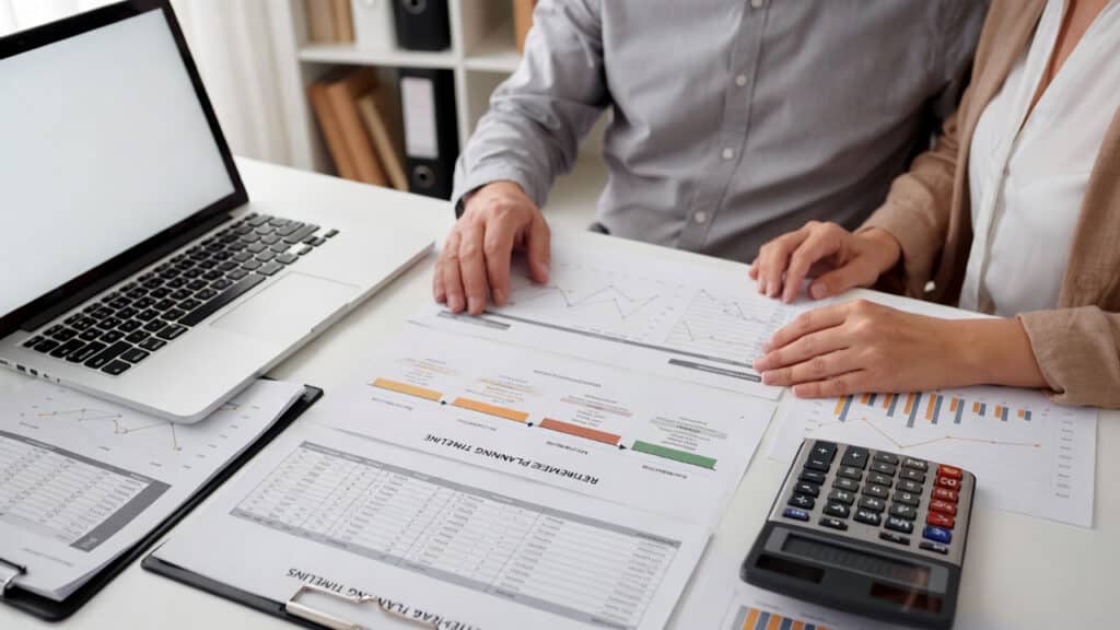 Two people sit at a desk reviewing financial charts and graphs on paper, with a laptop, calculator, and documents organized around them in an office setting.